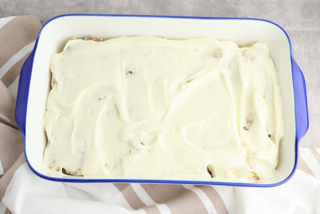 Cream cheese icing covering cinnamon roll pieces in baking dish.