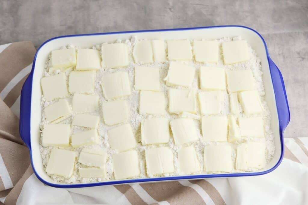 Butter pieces, coffee cake mix and cream cheese icing covering cinnamon roll pieces in baking dish.