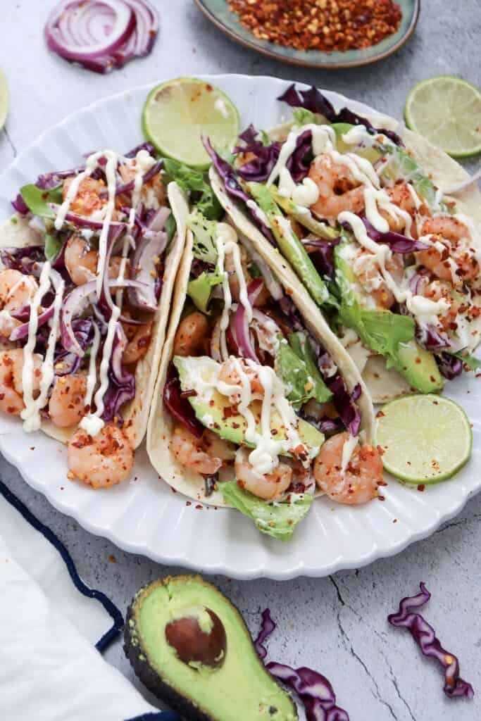 Spicy Shrimp Tacos on scalloped white plate, overhead shot, surrounded by avocados and red pepper flakes.