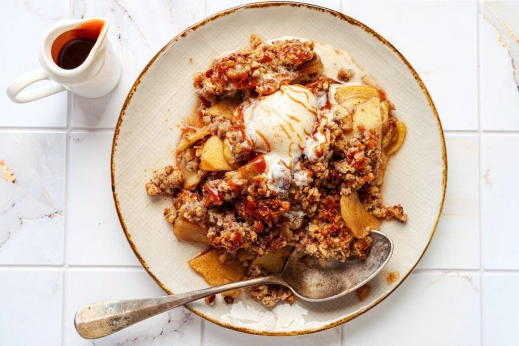 Apple Crisp with Vanilla Ice Cream on a plate with a spoon next to a ceramic dispenser of caramel sauce, overhead shot.