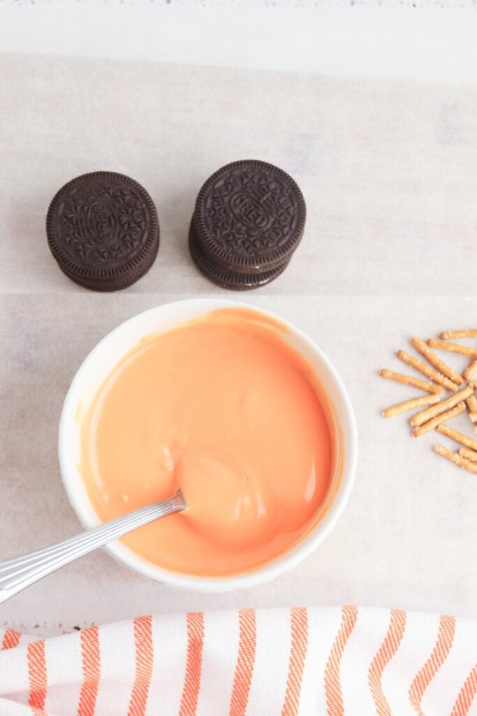 Oreo Pumpkins, in process, melted orange candy melt in bowl with spoon, and stacked Oreos, overhead shot.