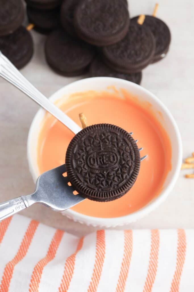 Oreo Pumpkins, in process, melted orange candy melt in bowl with Oreos ready to dip, held with forks.