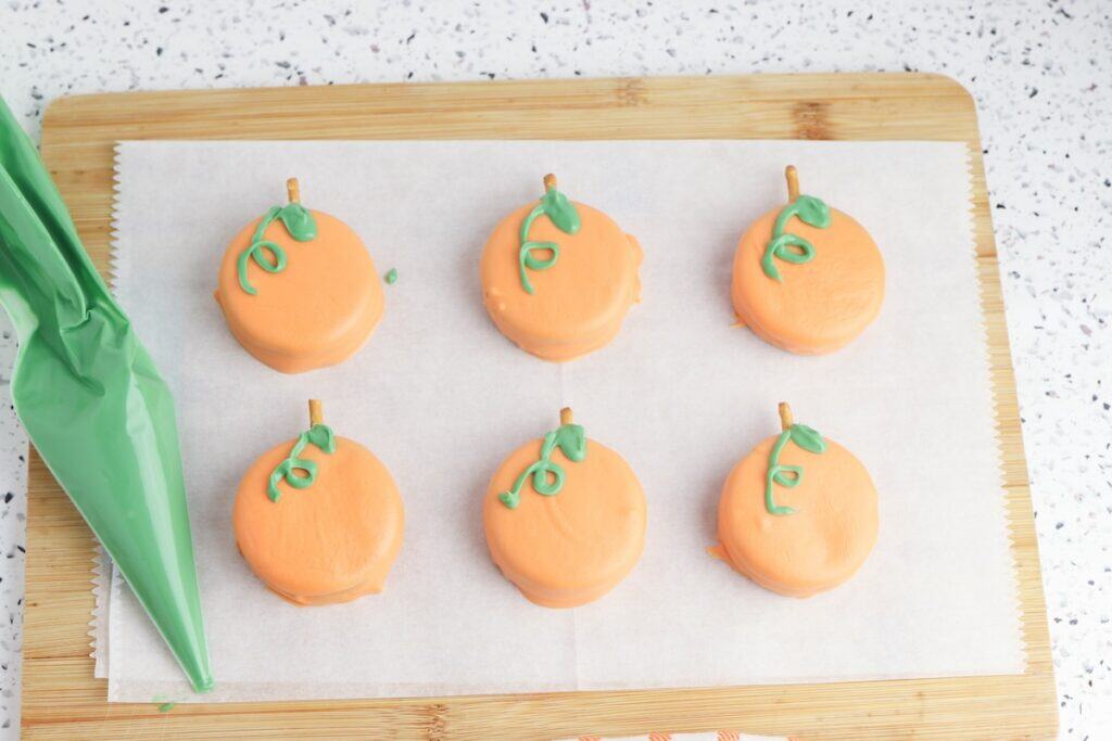 Oreo Pumpkins, in process, with green candy melt piping bag, on parchment paper on wooden cutting board.