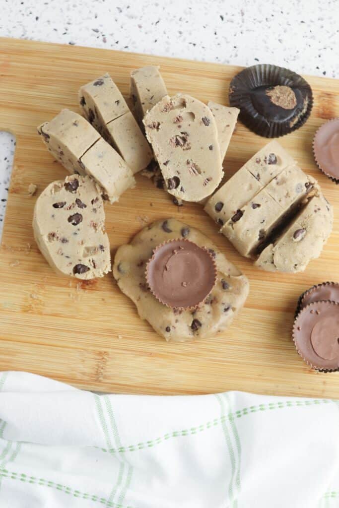 Reese’s Stuffed Cookies in process, chocolate chip cookie dough log sliced, with fun size Reese's Peanut Butter cups on cookie, on cutting board.