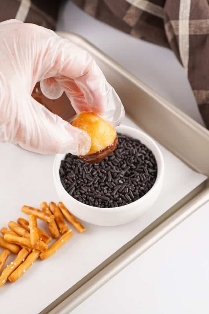 Acorn Donut Holes, in process, dipping chocolate-covered donut holes in chocolate sprinkles.