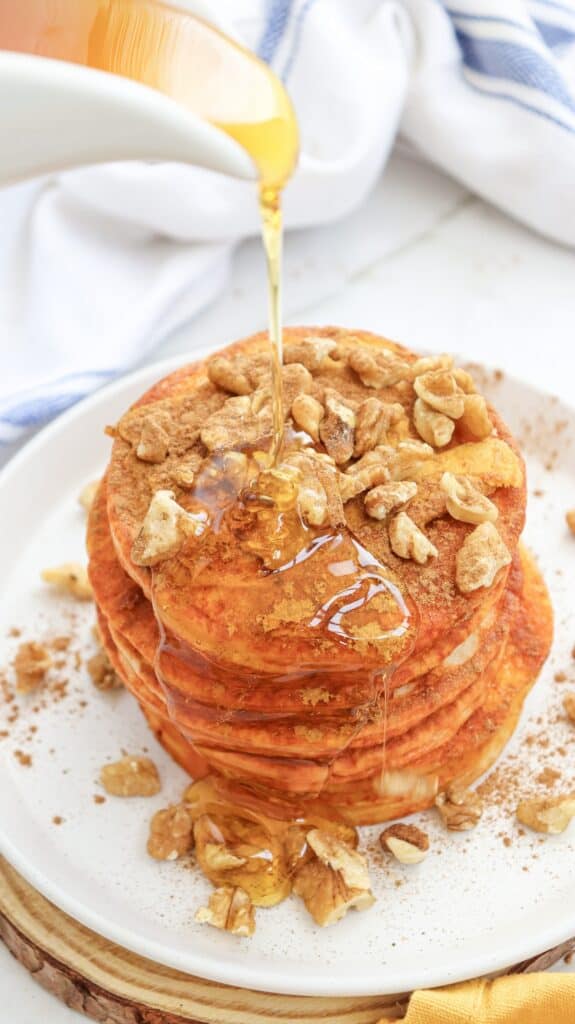 Stack of pumpkin pancakes covered in walnuts, with maple syrup being poured over the top.