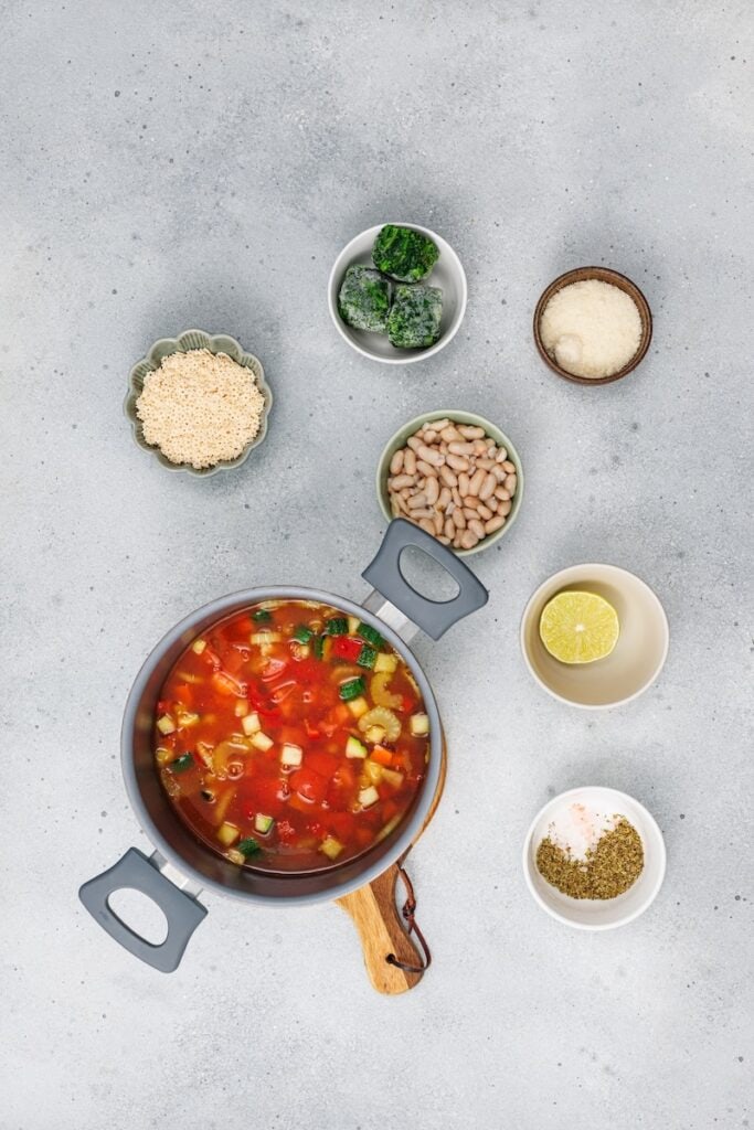 Veggie Minestrone Soup, in process, cooking tomatoes and vegetable broth in pot with other ingredients on the side.