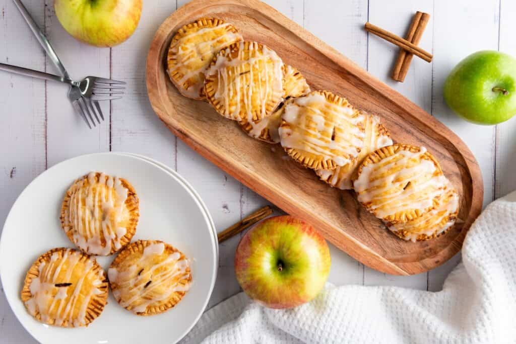 Apple Hand Pies with Flaky Crust and Sweet Cinnamon Filling on wooden serving tray and white plate, alongside apples, white wood background.