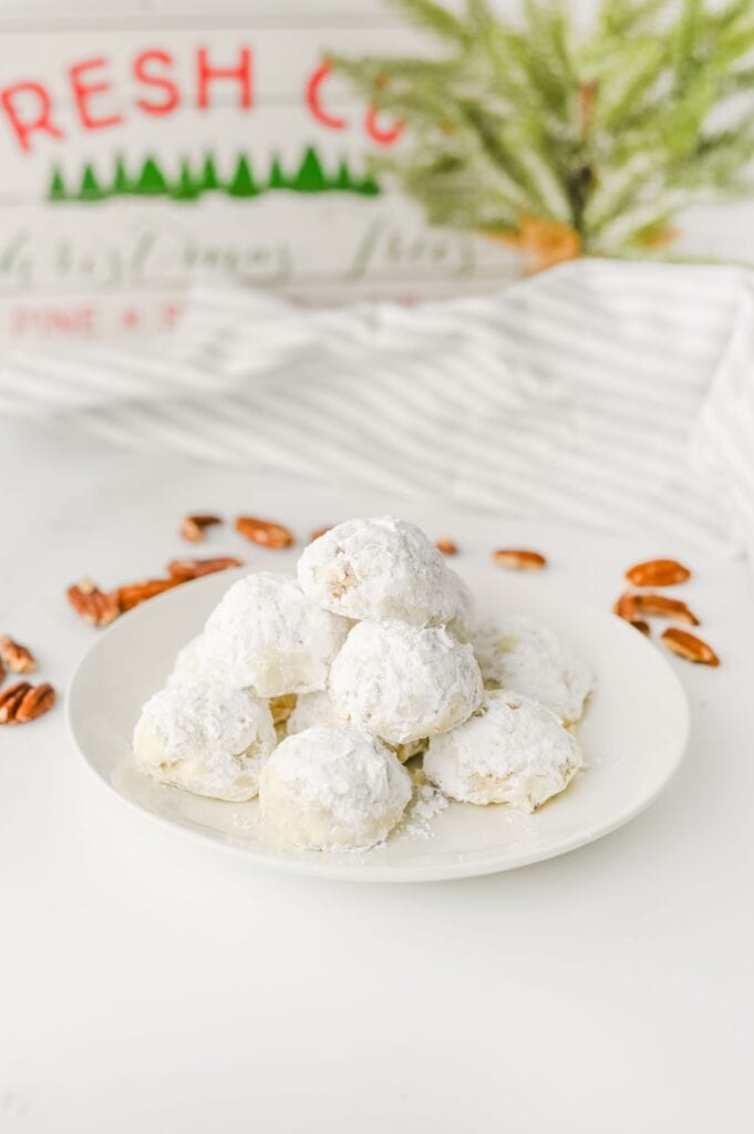 Pecan Snowball Cookies on white plate in kitchen surrounded by pecan pieces.