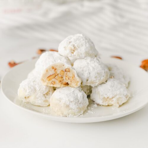 Pecan Snowball Cookies on white plate in kitchen surrounded by pecan pieces.